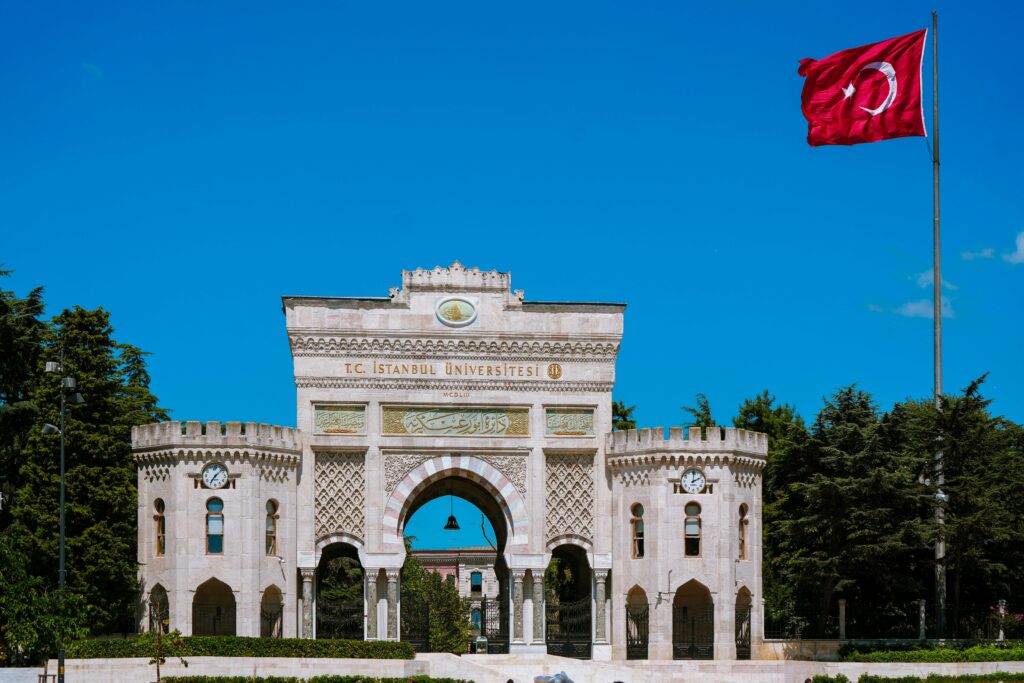 The grand entrance of Istanbul University adorned with a Turkish flag under a clear blue sky.