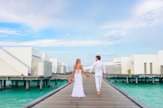 Full body back view of unrecognizable loving couple in white clothes walking on boardwalk above water near buildings while holding hands and looking at each other